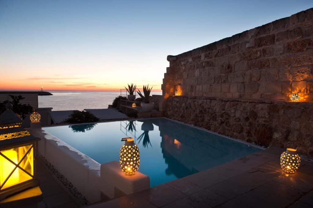 Parte do Hotel Don Ferrante que mostra a piscina ao ar livre com algumas luminárias acesas, alguns vasos de plantas e um muro de pedra. A vista da piscina é para o mar e para o céu durante o anoitecer. Imagem ilustrando post Hotéis em Puglia. 