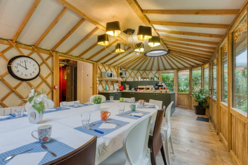 Sala de refeições do Hôtel du Jeu de Paume com uma mesa comprida posta com toalha, xícaras e copos. Alguns vasinhos de plantas decoram a mesa. Ao fundo há um balcão com micro-ondas e itens de café da manhã. Na parede ao lado esquerdo há um relógio, e todo o lado direito é composto de janelas com vista para a cidade.
