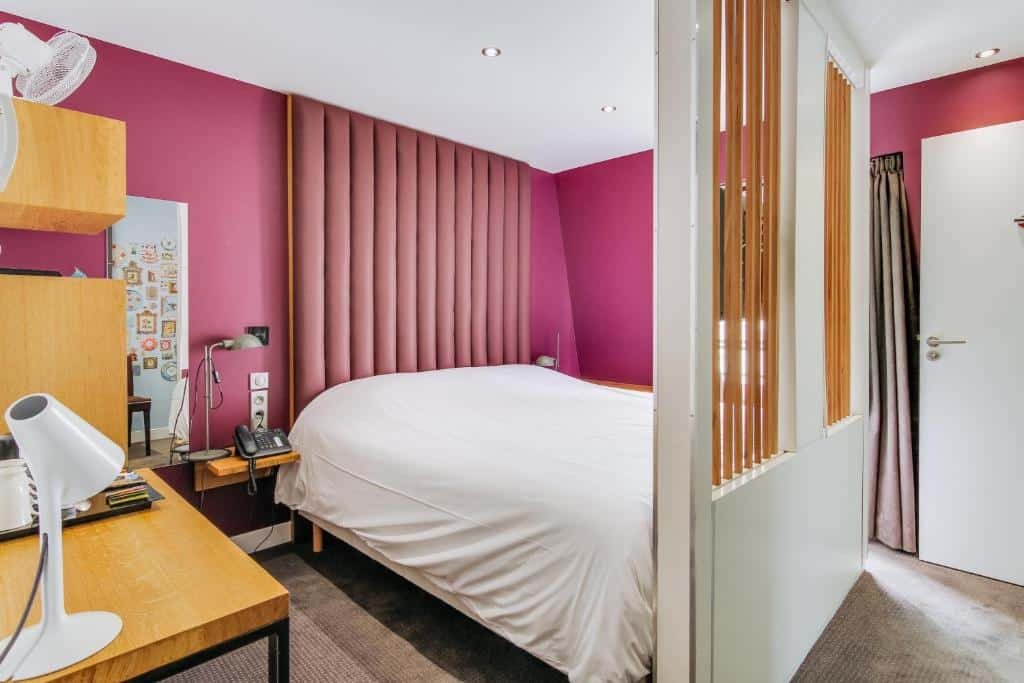 Quarto do Hôtel du Jeu de Paume, um dos hotéis perto do Palácio de Versalhes. Uma cama de casal está encostada na parede ao lado esquerdo com mesinhas de cabeceira e abajures dos dois lados, assim como um painel com TV logo aos pés. Na parede ao lado esquerdo fica uma mesa com abajur e comodidades para fazer chá e café. Já na parede ao lado direito estão a porta e a janela com cortinas.