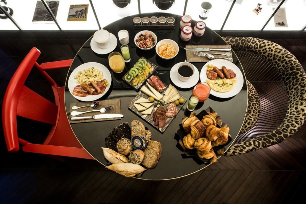 A imagem mostra uma mesa redonda preparada para um café da manhã farto no Hôtel du Rond-point des Champs-Élysées. Há pratos com ovos mexidos, bacon, salsichas, fatias de kiwi e manga, queijos e frios, além de pães variados e croissants. Também estão presentes bebidas como suco de laranja, suco verde e café. No centro da mesa, há tigelas com granola e iogurte. A mesa é rodeada por cadeiras modernas, incluindo uma vermelha e outra com estofado de estampa de oncinha. A iluminação suave e a apresentação organizada criam um ambiente sofisticado e convidativo.