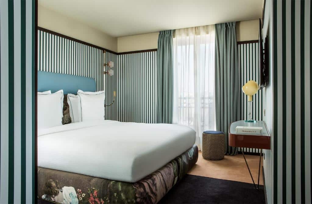A imagem mostra um quarto no Hôtel du Rond-point des Champs-Élysées, um dos hotéis com café da manhã em Paris. A cama é grande, com lençóis brancos impecáveis e uma cabeceira azul clara. As paredes são decoradas com listras verticais em verde e branco. Há uma janela grande com cortinas verdes claras que permitem a entrada de luz natural. Ao lado da cama, há um abajur moderno sobre uma mesa de cabeceira, junto a um pequeno banco redondo. O ambiente é acolhedor e bem iluminado, com uma decoração contemporânea e sofisticada.