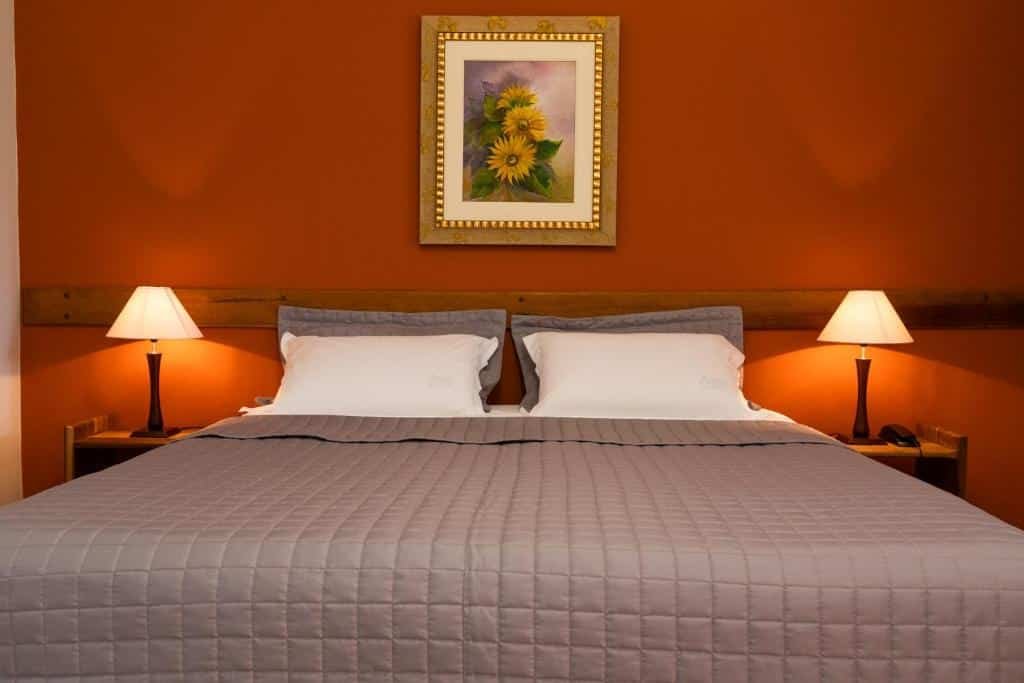 Quarto do Hotel Fazenda Areia que Canta com uma cama acolchoada cinza, dois abajures e uma pintura de flores amarelas em uma parede marrom-avermelhada.