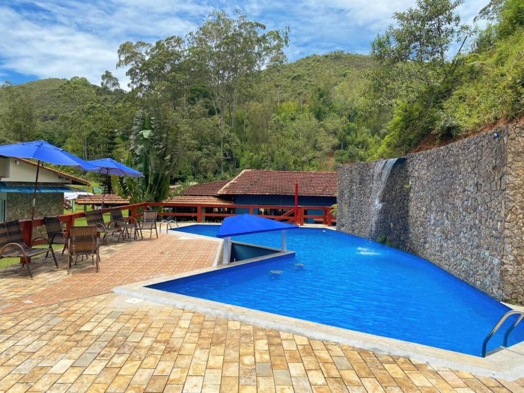 Área de piscina ao ar livre no Hotel Fazenda Bonanza com guarda-sóis azuis, espreguiçadeiras, uma cachoeira de pedra e vegetação ao redor em uma paisagem montanhosa.