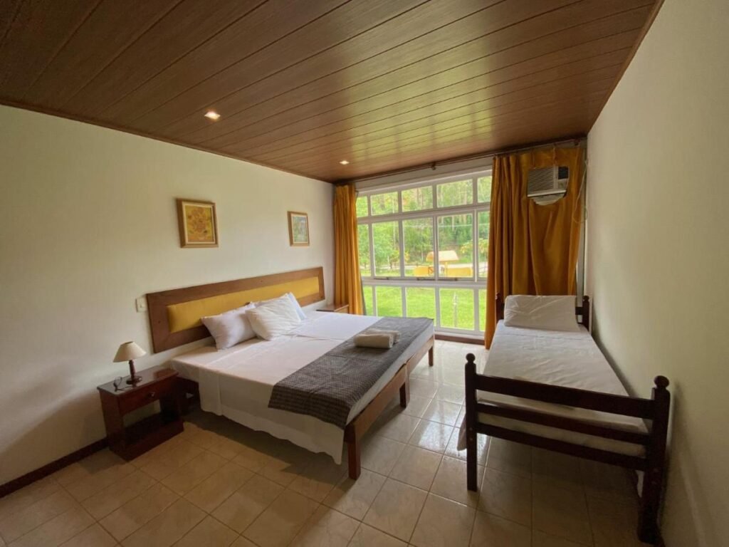Um quarto do Hotel Fazenda Bonanza com uma cama de casal e uma cama de solteiro, mesa de cabeceira com abajur, janela com cortinas e arte na parede. Esse é um dos hotéis fazenda no Rio de Janeiro.