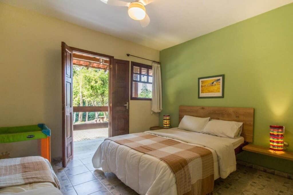 Um quarto do Hotel Fazenda Canto da Serra com uma cama de casal, mesas de cabeceira com luminárias, um ventilador de teto, uma parede verde e uma porta aberta para uma varanda.
