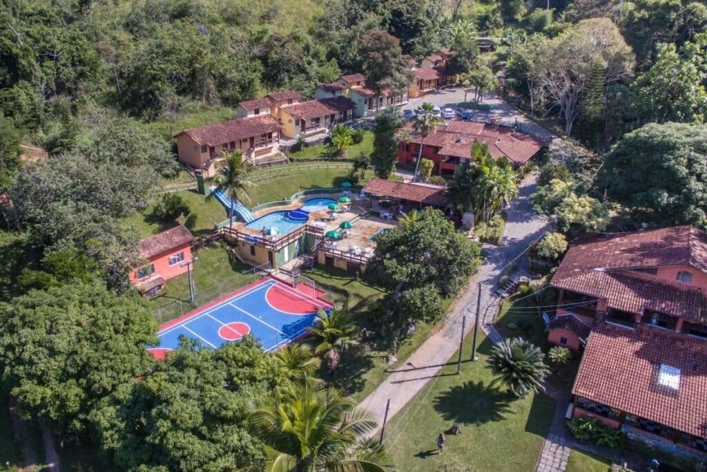 Vista aérea de uma área recreativa com piscina, quadra de basquete e vários prédios com telhados vermelhos cercados por árvores no Hotel Fazenda Canto da Serra, um dos hotéis fazenda no Rio de Janeiro.