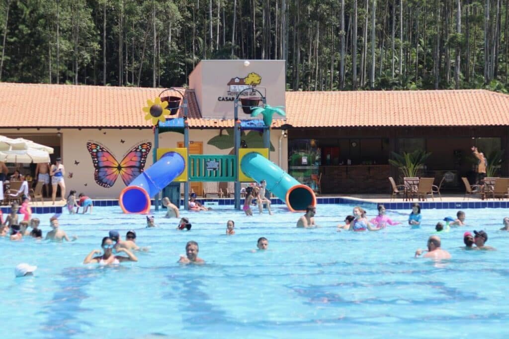 Pessoas de várias idades aproveitando uma grande piscina ao ar livre com toboáguas e um prédio ao fundo no Hotel Fazenda Casarão do Vale, um dos hotéis fazenda em Santa Catarina.