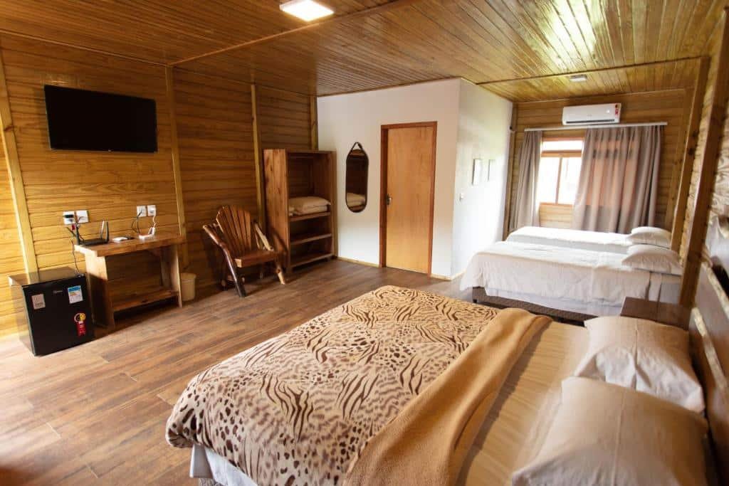 Um quarto de hotel mobiliado em madeira com duas camas, uma escrivaninha, uma TV, uma cadeira, uma geladeira pequena e uma janela com cortinasno Hotel Fazenda Casarão do Vale.