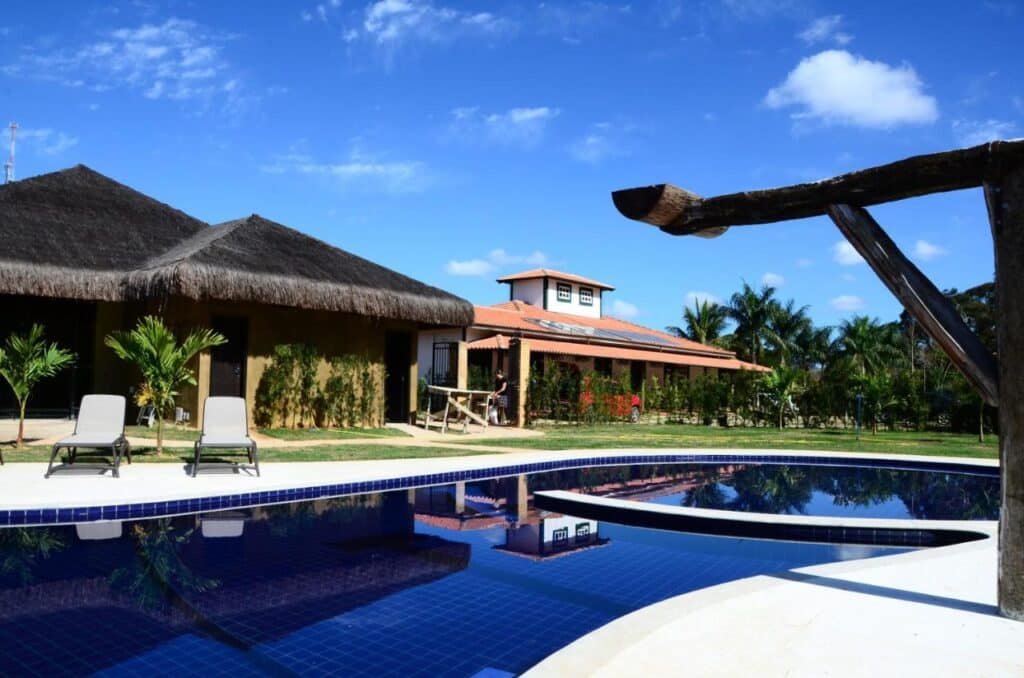 A imagem mostra uma área externa de um resort com uma piscina de azulejos azuis profundos. À esquerda, há duas espreguiçadeiras próximas à piscina, em frente a uma cabana com telhado de palha e paredes decoradas com plantas trepadeiras. Ao fundo, há uma construção maior com telhado de telhas vermelhas e varanda, cercada por jardins bem cuidados. O céu está azul com algumas nuvens, e várias palmeiras completam o ambiente tropical e relaxante. O clima parece ensolarado, ideal para desfrutar a piscina e o espaço ao ar livre. Esse é o Hotel Fazenda Cheiro Verde, um dos hotéis-fazenda em Minas Gerais.