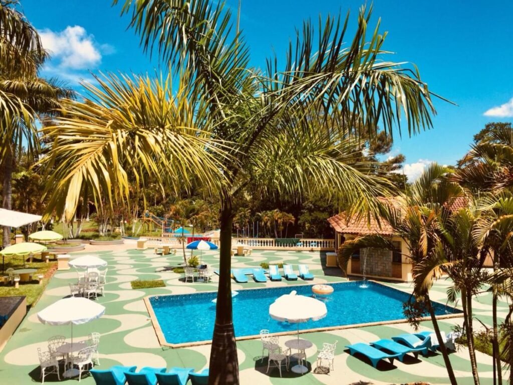 A imagem mostra uma área de lazer externa com uma piscina retangular cercada por espreguiçadeiras azuis e mesas brancas com guarda-sóis. Ao redor, há várias palmeiras e vegetação tropical, criando uma atmosfera de resort. O piso ao redor da piscina tem um design colorido em verde e cinza, formando um padrão geométrico. No fundo, há um pequeno edifício com telhado de cerâmica, e ao longe, é possível ver um parquinho infantil. O céu está azul com algumas nuvens, reforçando o clima ensolarado e relaxante do lugar. Esse é o Hotel Fazenda Colina, um dos hotéis-fazenda em Minas Gerais.