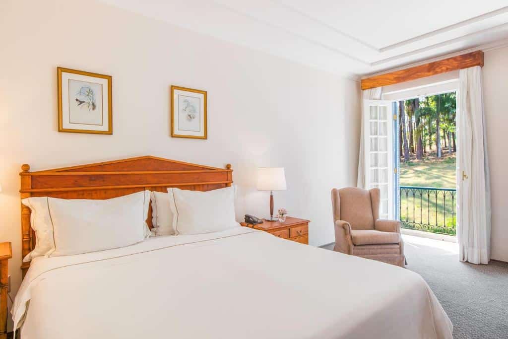 Um quarto do Hotel Fazenda Dona Carolina, em São Paulo, iluminado com uma cama grande, abajur, poltrona e portas abertas que levam a uma varanda com vista para as árvores.