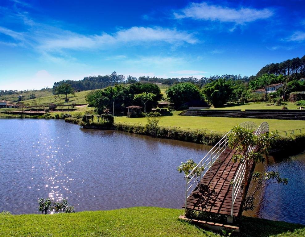 Uma pequena ponte de madeira em arco cruza um lago calmo no Hotel Fazenda Dona Carolina, com gramados verdes e árvores sob um céu azul claro ao fundo.