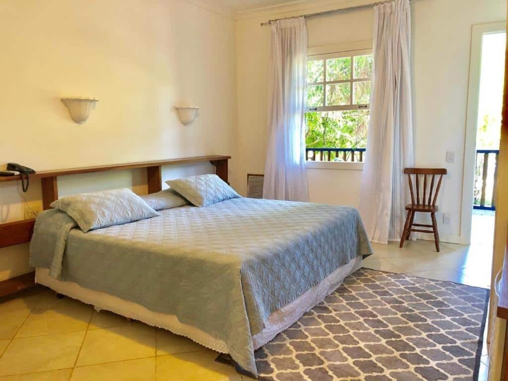 Um quarto do Hotel Fazenda Florença com uma cama grande, colcha azul, cabeceira de madeira, duas luminárias de parede, janela com cortinas e uma cadeira de madeira.