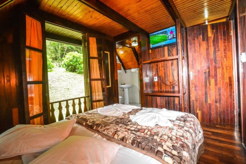 Um quarto aconchegante de cabine com painéis de madeira, uma cama de casal, TV e janelas abertas mostrando a vegetação lá fora. Uma pia é visível em um pequeno recanto. O local fica no Hotel Fazenda Floresta Negra, um dos hotéis fazenda em Minas Gerais.