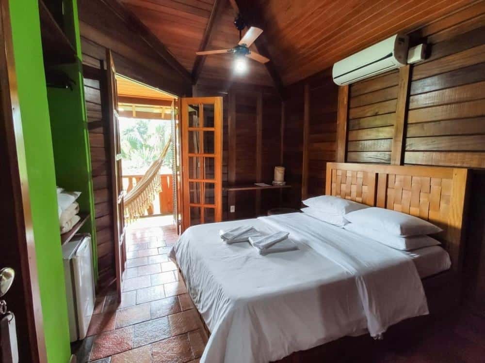 Um quarto de madeira com uma cama feita, ar condicionado e uma porta aberta que leva a uma varanda com rede no Hotel Fazenda Igarapés, um dos hotéis fazenda em Minas Gerais.
