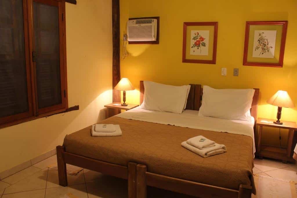 Um quarto do Hotel Fazenda Jacaúna aconchegante com uma cama de casal, duas mesas de cabeceira com luminárias, ar condicionado e uma obra de arte emoldurada em uma parede amarela.