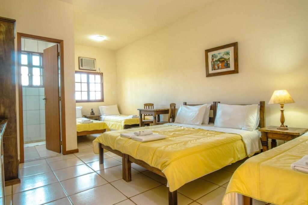 A imagem mostra um quarto simples e bem iluminado do Hotel Fazenda Minas Real, com três camas de solteiro cobertas com colchas amarelas. Ao lado de uma das camas, há um criado-mudo de madeira com um abajur aceso, proporcionando uma luz suave. O chão é de azulejo branco e há uma janela com moldura de madeira, permitindo a entrada de luz natural. Na parede, há um quadro decorativo. Ao fundo, é possível ver a porta aberta para o banheiro, e há uma pequena mesa com cadeiras de madeira perto da janela. O ambiente é acolhedor e organizado.