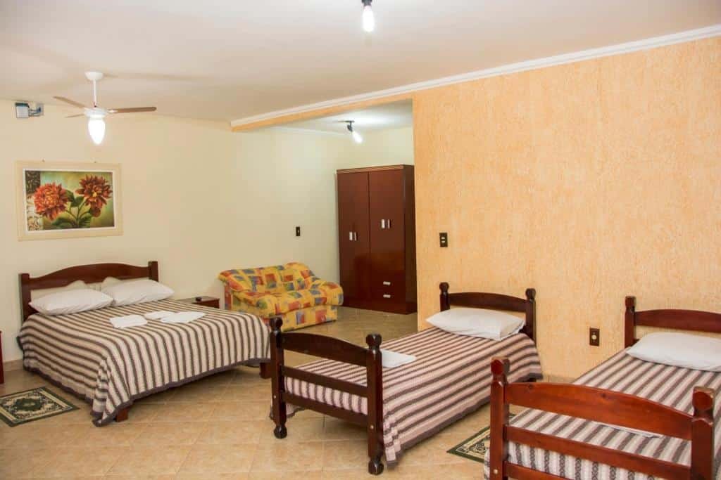 A imagem mostra um quarto espaçoso do Hotel Fazenda Monte Sião com três camas, uma de casal e duas de solteiro, todas com colchas listradas em tons de marrom e branco. As camas têm cabeceiras de madeira, e o piso é de cerâmica bege. Ao fundo, há um armário marrom de duas portas e um sofá com estampa colorida em amarelo e laranja. Uma parede laranja clara ao lado contrasta com as outras paredes brancas. Há um ventilador de teto e uma luminária simples no centro do quarto, além de um quadro com flores coloridas na parede. O ambiente é simples, mas acolhedor.