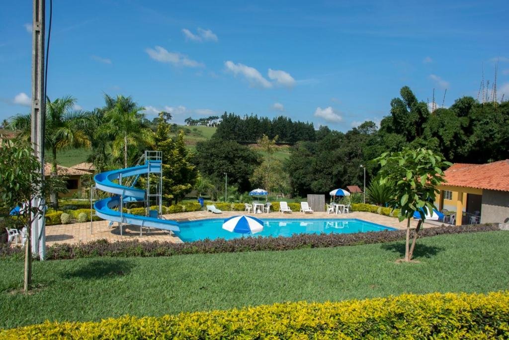A imagem mostra uma área de lazer ao ar livre com uma piscina retangular cercada por cadeiras e mesas brancas com guarda-sóis azuis e brancos. Ao lado da piscina, há um tobogã em espiral de cor azul, adicionando um elemento divertido ao local. O ambiente é cercado por gramado bem cuidado e arbustos, com palmeiras e outras árvores ao redor. Ao fundo, é possível ver colinas verdes e uma densa vegetação, criando uma atmosfera tranquila e natural. O céu está claro e azul, sugerindo um dia ensolarado. Esse é o Hotel Fazenda Monte Sião, um dos hotéis-fazenda em Minas Gerais.