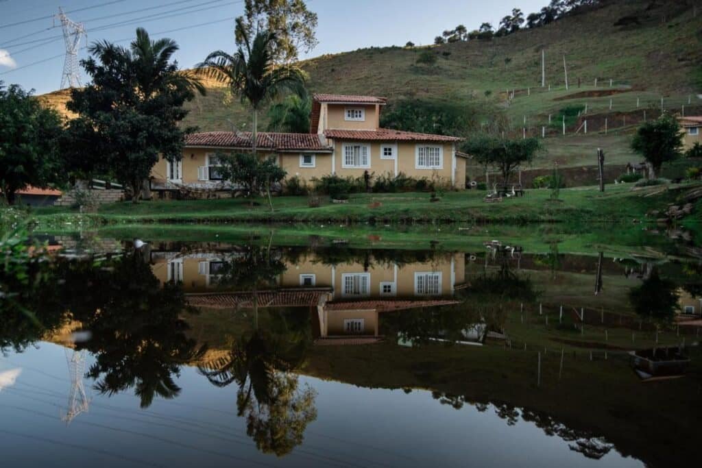 Uma casa com telhado de telhas vermelhas é refletida em um lago calmo, cercada por grama, árvores e uma encosta ao fundo. Esse é o Hotel Fazenda Monteiro, um dos hotéis fazenda no Rio de Janeiro.
