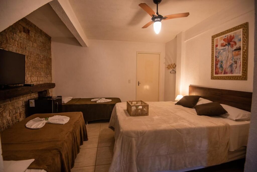 Um quarto do Hotel Fazenda Monteiro com uma cama grande, uma cama de solteiro, uma TV sobre uma prateleira de pedra, um ventilador de teto e um quadro emoldurado na parede.