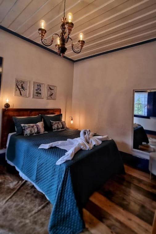 Um pequeno quarto aconchegante com uma colcha azul-petróleo, esculturas de cisnes em toalhas na cama, arte na parede acima da cabeceira e um lustre. O local fica no Hotel Fazenda Paciência, um dos hotéis fazenda em Minas Gerais.