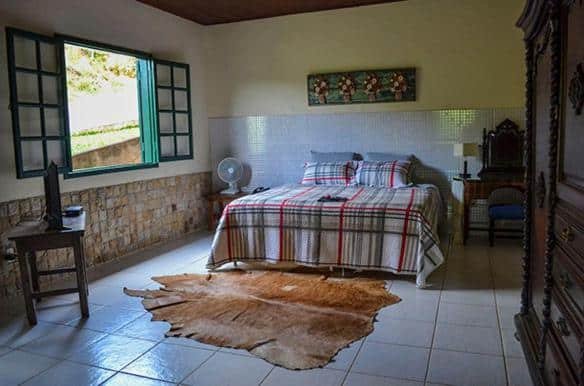 Um quarto aconchegante com uma cama de casal, janela aberta, móveis de madeira, um ventilador sobre a mesa e um tapete de couro no chão de ladrilhos no Hotel Fazenda Pirapetinga.