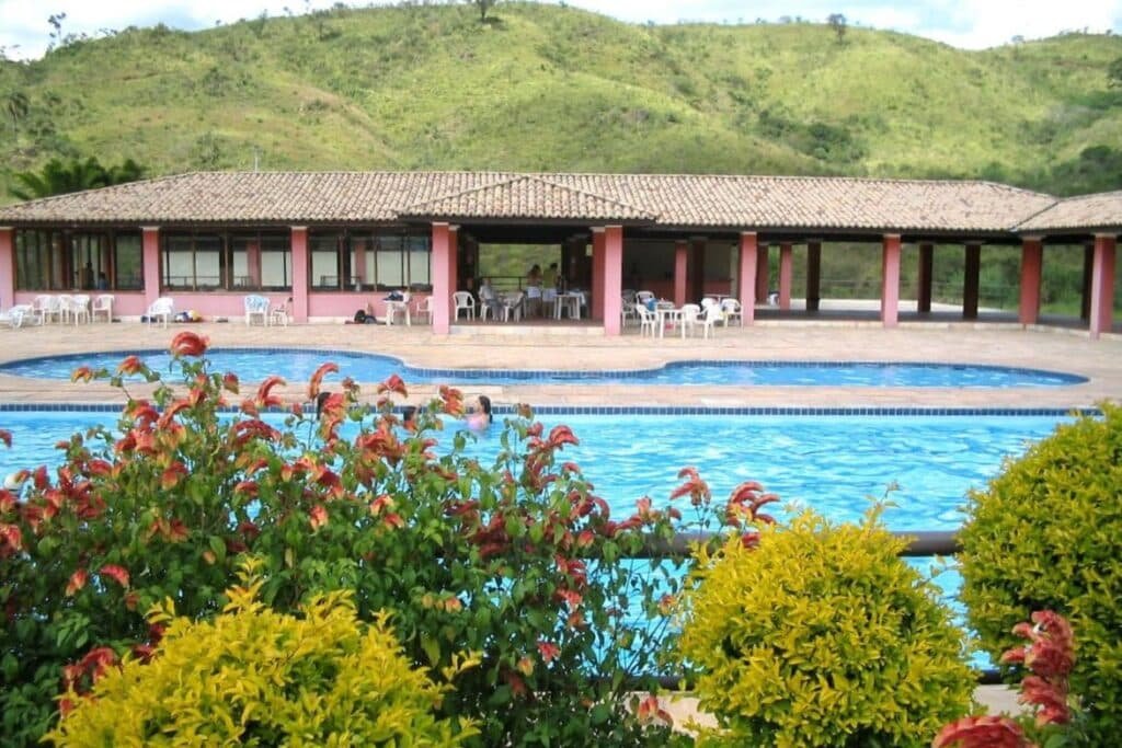 Área de piscina ao ar livre do Hotel Fazenda Retiro das Rosas com guarda-sóis, mesas e cadeiras. Colinas e vegetação ao fundo, com arbustos floridos coloridos em primeiro plano.