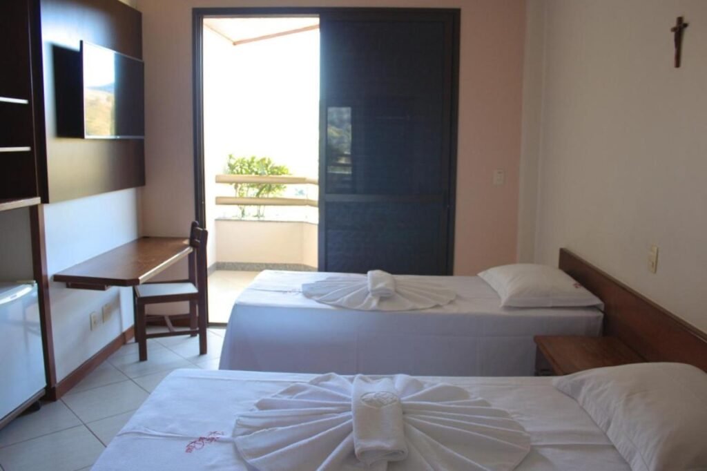 Um quarto de hotel com duas camas de solteiro, uma escrivaninha, uma TV, uma mini geladeira e uma porta de sacada. Toalhas são dobradas decorativamente nas camas desse local que fica no Hotel Fazenda Retiro das Rosas, um dos hotéis fazenda em Minas Gerais.