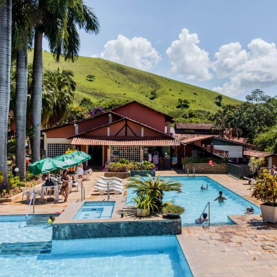 Área de piscina ao ar livre no Hotel Fazenda Rochedo, um dos hotéis fazenda no Rio de Janeiro, com pessoas relaxando e nadando, cercada por palmeiras e uma encosta ao fundo.