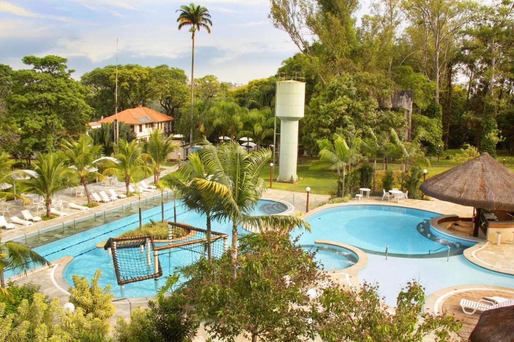 Uma área de piscina do Hotel Fazenda Salto Grande em São Paulo com uma ponte, espreguiçadeiras e um bar com teto de palha, cercada por palmeiras e vegetação exuberante.