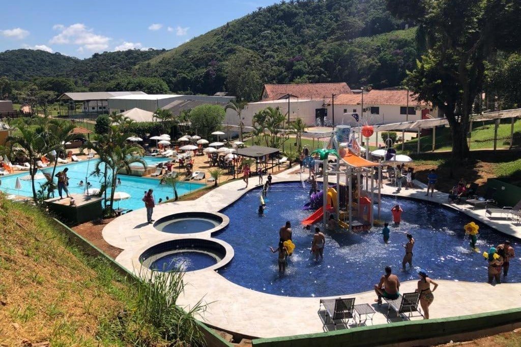 Vista de um parque aquático no Hotel Fazenda Santa Barbara com pessoas nadando em piscinas, brincando em toboáguas e relaxando sob guarda-sóis cercados por vegetação.