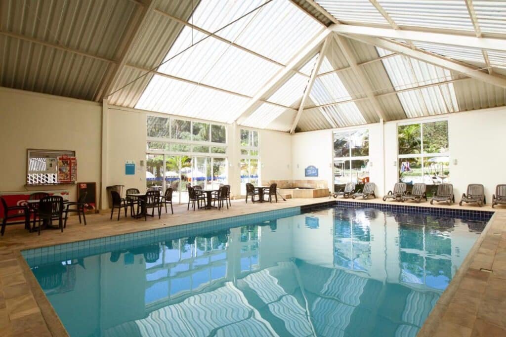 Piscina coberta com água limpa, cercada por cadeiras no Hotel Fazenda Santa Barbara, um dos hotéis fazenda no Rio de Janeiro. Grandes janelas e um teto alto deixam entrar luz natural.