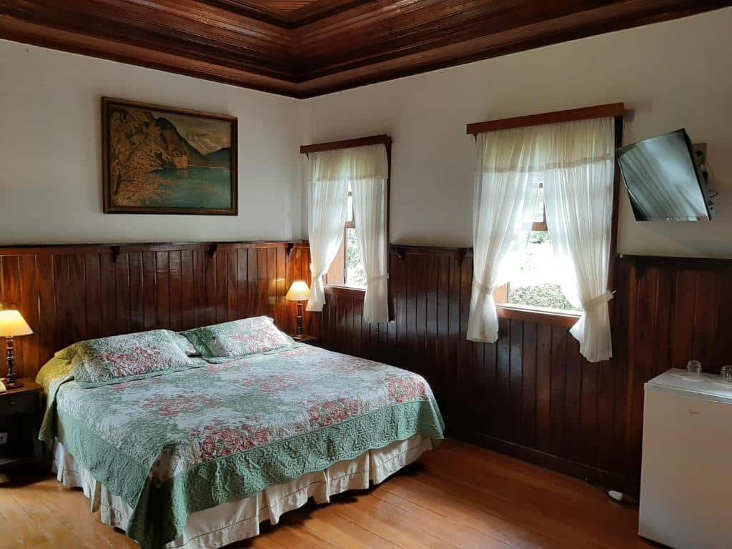 Um quarto aconchegante no Hotel Fazenda São Moritz com painéis de madeira, uma cama de casal com roupa de cama floral, duas luminárias, uma TV montada na parede e duas janelas com cortinas.