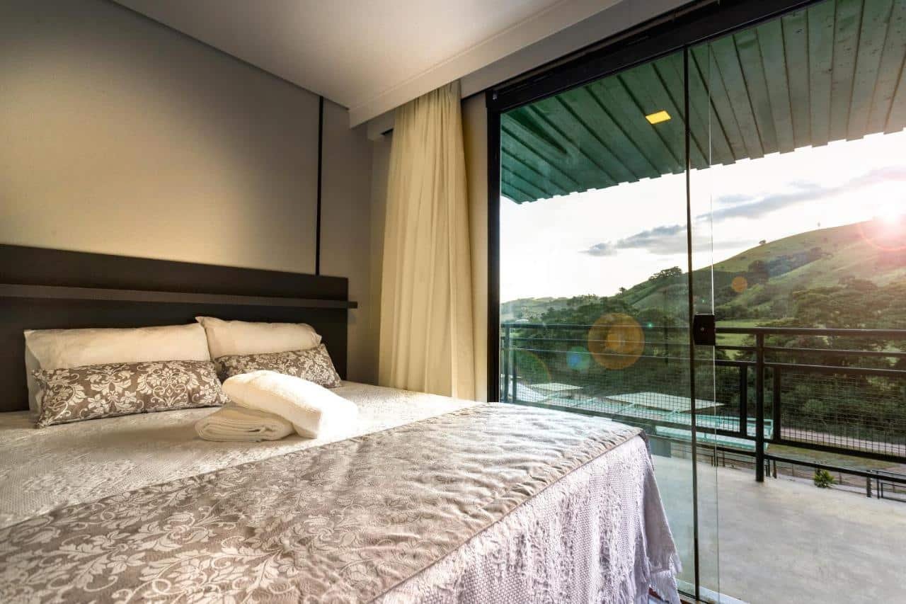 Um quarto com uma cama grande, roupa de cama branca e cinza, e toalha branca. Portas de vidro abrem para uma sacada com vista para colinas verdes. Esse quarto é do Hotel Fazenda Terra dos Sonhos, um dos hotéis fazenda em Minas Gerais.