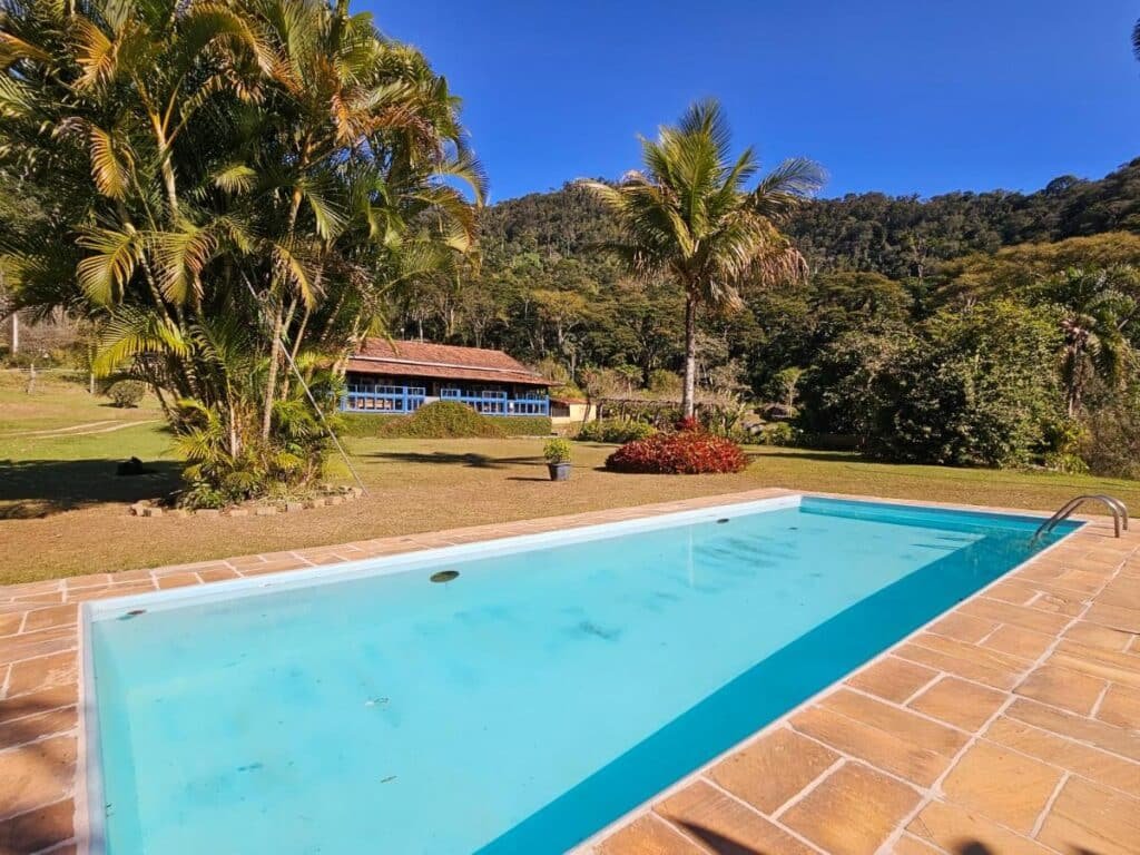Uma piscina retangular com água limpa é cercada por um deck de pedra, com um gramado verde, palmeiras e um edifício azul ao fundo no Hotel Fazenda Upã Moña, um dos hotéis fazenda no Rio de Janeiro.