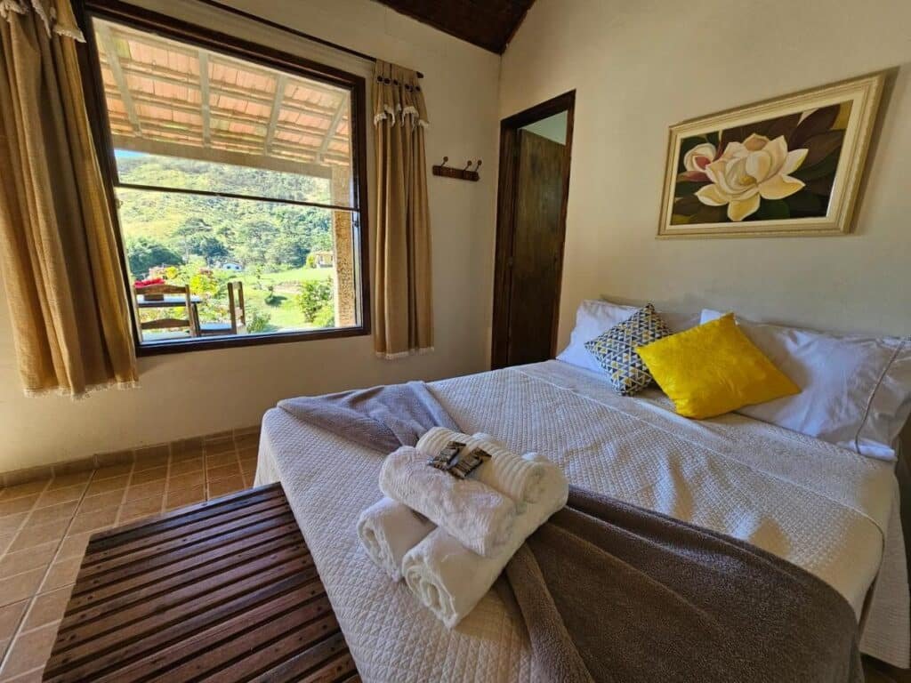 Um quarto aconchegante no Hotel Fazenda Upã Moña com uma cama bem arrumada, toalhas dobradas em cima, um banco de madeira, uma grande janela com vista panorâmica e obras de arte florais.