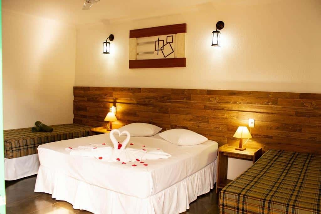 A imagem mostra um quarto de hotel aconchegante com uma cama de casal central e duas camas de solteiro nas laterais. A cama de casal está decorada com toalhas dobradas em forma de cisnes e pétalas vermelhas, criando um clima romântico. As paredes têm uma iluminação suave, com luminárias de estilo rústico sobre a cabeceira de madeira. Ao lado da cama, há pequenos abajures sobre mesas de cabeceira, que complementam a atmosfera acolhedora e convidativa do quarto. O ambiente é simples, mas bem arrumado e iluminado, ideal para descanso. Esse é o Hotel Fazenda Vale Amanhecer, um dos hotéis-fazenda em Minas Gerais.