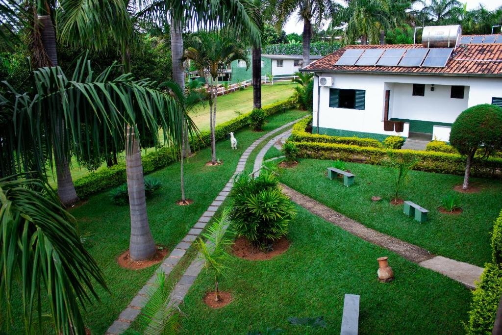 A imagem mostra um jardim bem cuidado, com grama verde e caminhos de pedras que serpenteiam pelo espaço no Hotel Fazenda Vale Amanhecer. Há várias palmeiras altas e plantas ornamentais ao redor, além de bancos verdes de madeira posicionados em áreas estratégicas para descanso. No canto direito, uma construção branca com janelas verdes e telhado de telhas vermelhas é visível, com painéis solares no telhado. O cenário é tranquilo, com um ambiente natural e organizado, ideal para passeios e momentos de relaxamento ao ar livre.