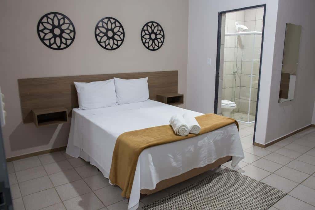 Um quarto do Hotel Fazenda Vale da Cachoeira bem arrumado com uma cama, dois travesseiros, toalhas dobradas, arte decorativa na parede e uma porta de banheiro aberta.