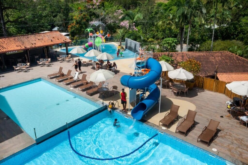 Vista aérea de uma área de piscina do Hotel Fazenda Vilarejo All Inclusive com toboáguas, espreguiçadeiras, guarda-sóis e pessoas nadando e brincando perto de um playground aquático.