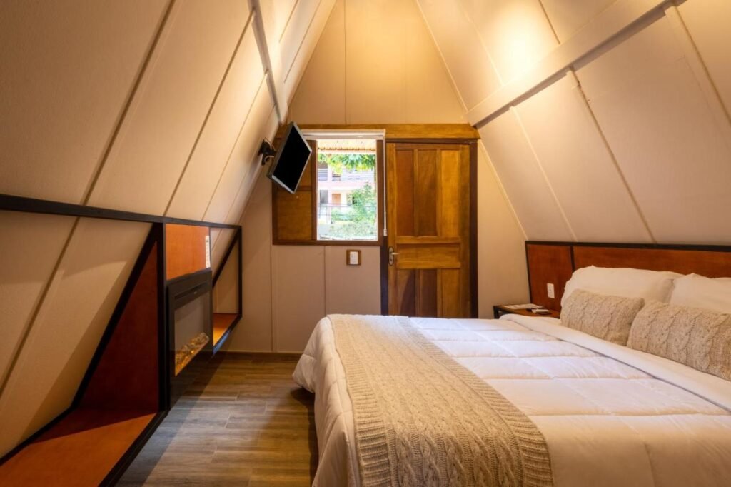 Um quarto aconchegante em formato de A com porta de madeira, cama com lençóis brancos, TV montada na parede e uma pequena lareira elétrica no Hotel Fazenda Vilarejo All Inclusive, um dos hotéis fazenda no Rio de Janeiro.