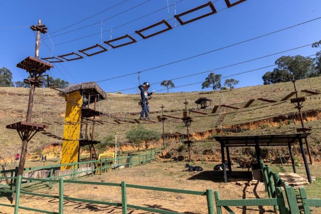Pessoa praticando arvorismo em um percurso de cordas altas com plataformas e escadas, em uma área externa montanhosa com terreno gramado no Hotel Fazenda Vilarejo All Inclusive.