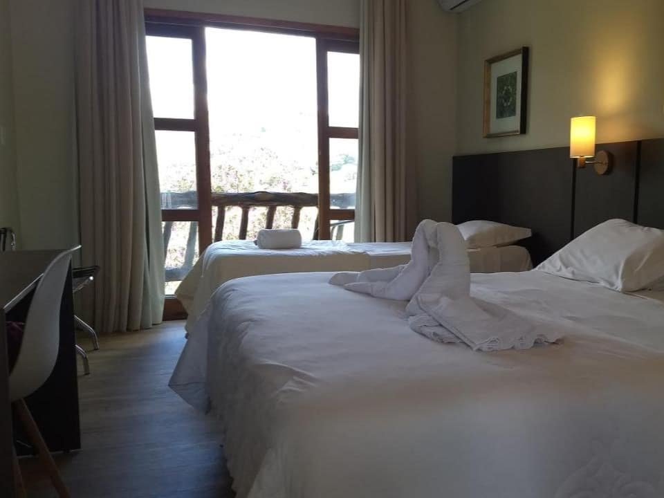 Um quarto do Hotel Fazenda Vista Alegre com duas camas, uma escrivaninha de madeira com uma cadeira, uma grande janela com cortinas e uma toalha dobrada em forma de cisne em uma das camas. Esse é um dos hotéis fazenda em Minas Gerais.