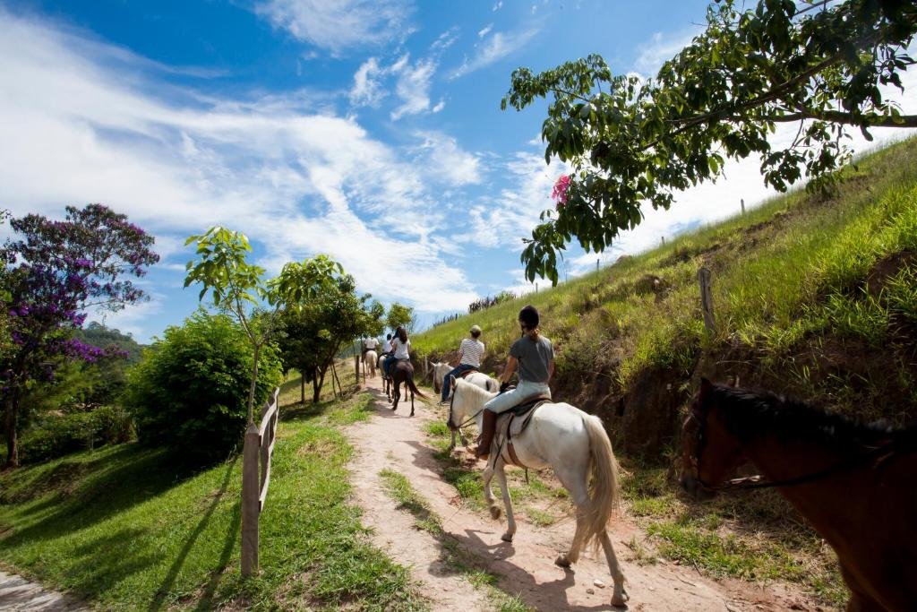 Várias pessoas andando a cavalo em uma trilha de terra com muito verde e árvores ao redor, sob um céu azul com nuvens dispersas.