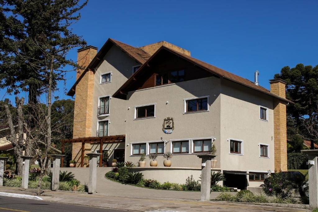 vista frontal do Hotel Gramado Interlaken mostrando a entrada de carro com uma rampa para o lounge do hotel. É uma construção de três andares e ao seu lado há algumas araucárias