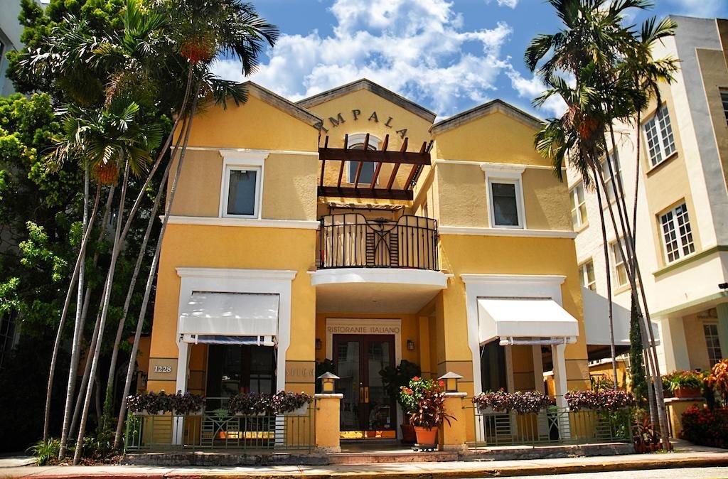A imagem mostra a fachada de um prédio de estilo mediterrâneo pintado de amarelo claro, com detalhes brancos. O prédio tem dois andares e um pequeno balcão com uma estrutura de madeira no segundo andar. A entrada principal tem toldos brancos e é cercada por plantas e flores em vasos. Há palmeiras altas ao lado do prédio, adicionando um toque tropical. O nome "Impala" está visível no topo da fachada. O céu ao fundo é azul com algumas nuvens, criando um cenário agradável e ensolarado. O local é um dos hotéis em Miami Beach indicados.