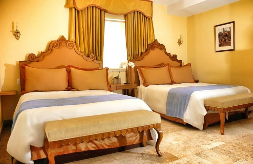 A imagem mostra um quarto do Impala Hotel decorado em estilo clássico com duas camas de casal. As camas têm cabeceiras ornamentadas de madeira e são cobertas com roupa de cama branca, travesseiros bege e colchas azul claro. Cada cama possui um banco estofado aos pés. As paredes são amarelas, combinando com as cortinas douradas que enquadram uma janela entre as camas. No centro, há uma mesa de cabeceira com flores brancas. A decoração é complementada por uma luminária de parede e um quadro na parede lateral, criando uma atmosfera acolhedora e elegante.