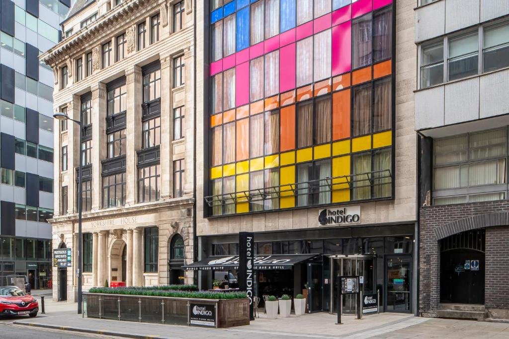 Frente do Hotel Indigo Liverpool, com andares coloridos (sendo o primeiro amarelo, o segundo laranja, o terceiro rosa e o parte de um andar azul), além de prédios ao lado na mesma rua