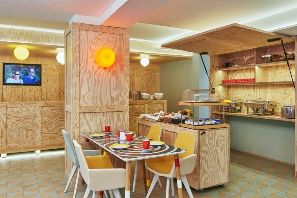 A imagem mostra uma área de café da manhã com decoração moderna e colorida no Hotel Joke - Astotel. Há uma mesa com quatro cadeiras de cores diferentes (amarela, branca e cinza) e pratos e canecas vermelhas. Ao fundo, há um buffet com diversos itens de café da manhã, como pães, frutas e cereais, dispostos em prateleiras e recipientes de madeira clara. As paredes e colunas são revestidas de madeira com um acabamento rústico. Há também uma televisão na parede e luzes decorativas coloridas, criando um ambiente acolhedor e vibrante.